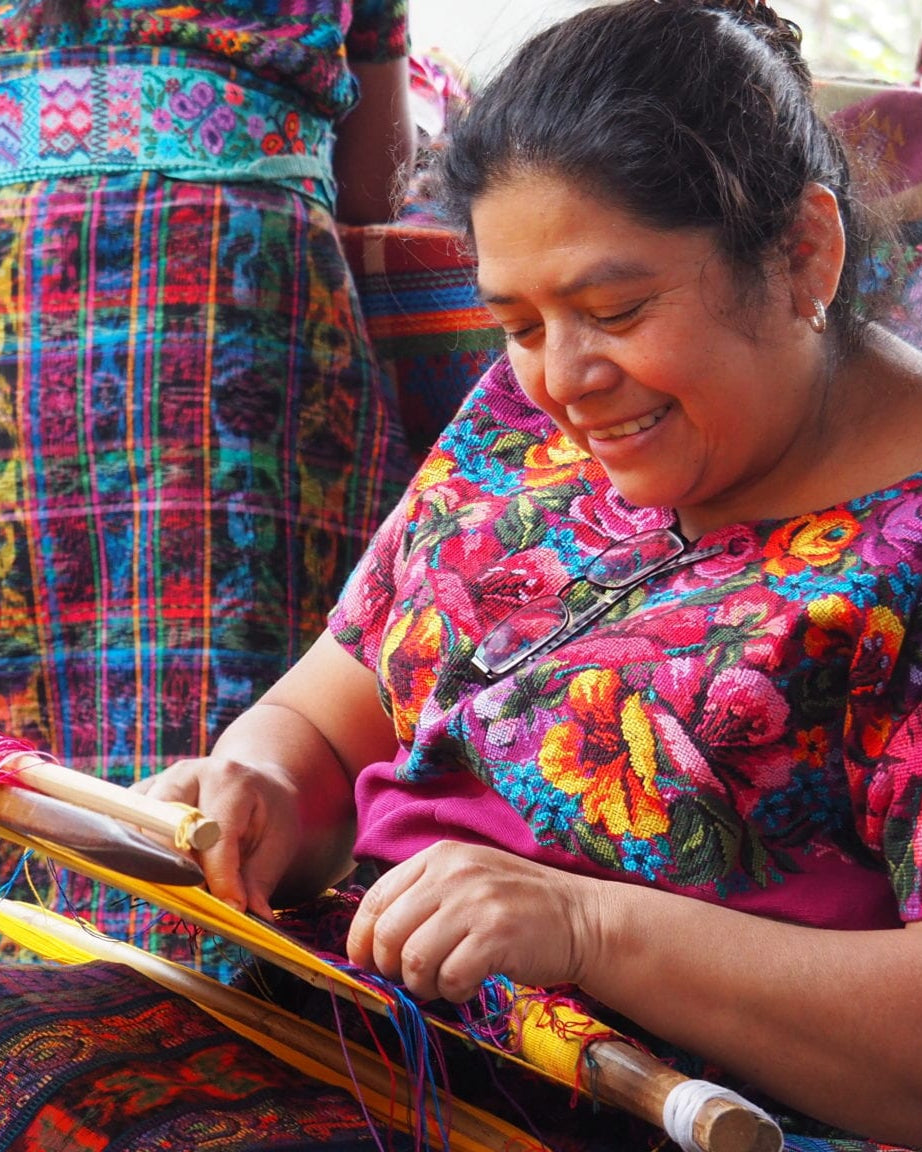 Woman in colorful traditional clothing using a loom with vibrant textiles in the background.